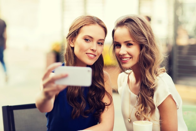 tecnologia, estilo de vida, amizade e conceito de pessoas - mulheres jovens ou adolescentes felizes com smartphone e xícaras de café tomando selfie no café ao ar livre