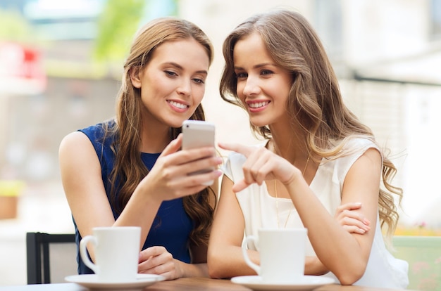 tecnología, estilo de vida, amistad y concepto de personas: mujeres jóvenes felices o adolescentes con teléfonos inteligentes y tazas de café en un café al aire libre