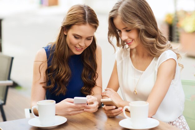 tecnología, estilo de vida, amistad y concepto de personas: mujeres jóvenes felices o adolescentes con teléfonos inteligentes y tazas de café en el café al aire libre