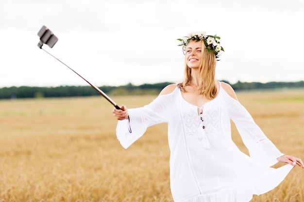 tecnologia, férias de verão, férias e conceito de pessoas - jovem sorridente em coroa de flores tirando fotos com bastão de selfie de smartphone no campo de cereais