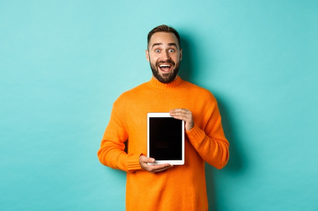 Tecnologia. Homem adulto empolgado com suéter laranja mostrando a tela do tablet digital, olhando surpreso para