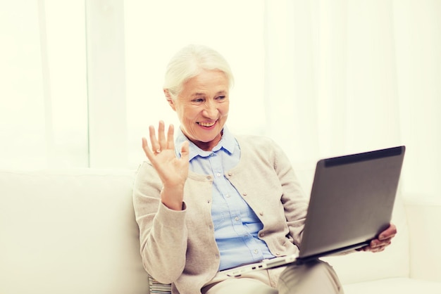 tecnologia, idade, gesto, comunicação e conceito de pessoas - mulher sênior feliz com computador portátil tendo chat de vídeo em casa e acenando com a mão