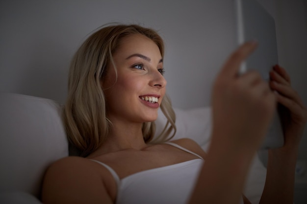 tecnología, internet, comunicación y concepto de personas - mujer joven sonriente feliz con computadora de tablet pc en la cama en el dormitorio de casa por la noche