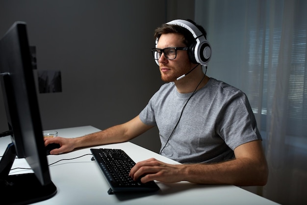 tecnología, juegos, entretenimiento, juguemos y concepto de personas: joven con auriculares y anteojos con computadora de PC jugando en casa y transmitiendo videos de recorrido o recorrido