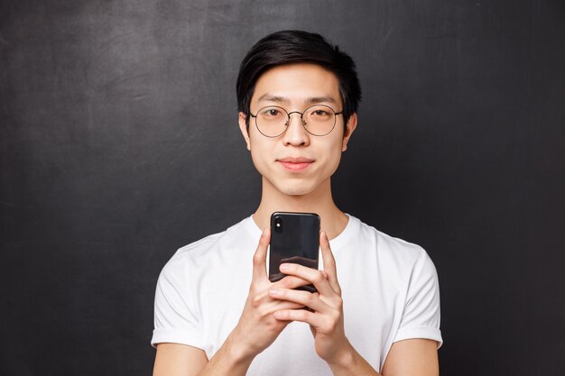 Tecnología, mensajería y concepto de personas. Retrato de primer plano de joven estudiante masculino asiático sonriendo, sosteniendo el teléfono inteligente y mirando la cámara, enviando mensajes de texto a un amigo, usando la aplicación,