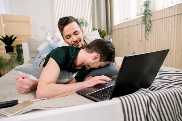 Foto tecnología moderna. bastante joven pareja gay usando laptop mientras posa en la cama y pasar tiempo juntos