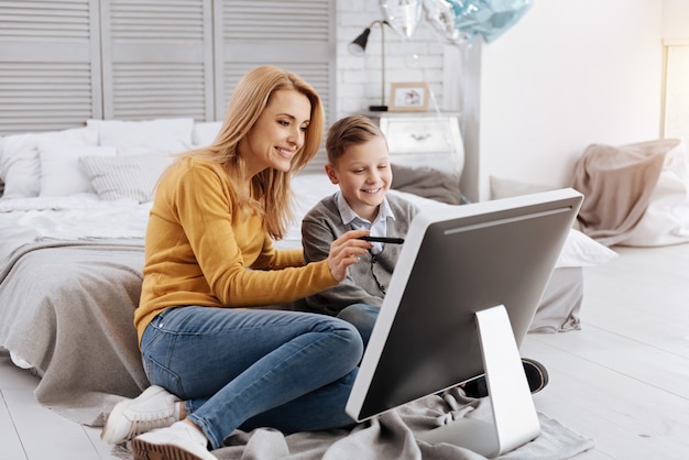 Tecnología progresiva. Encantado de mujer joven positiva sentada junto a su hijo y sonriendo mientras le muestra la nueva tecnología