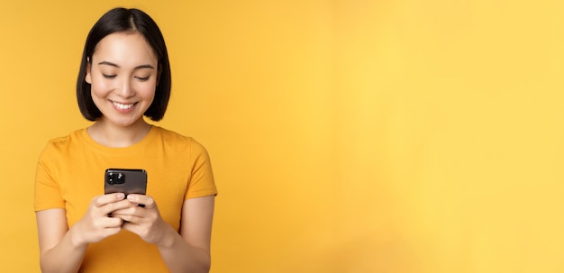 Tecnología sonriente mujer asiática usando teléfono móvil con teléfono inteligente en las manos de pie en camiseta aga...