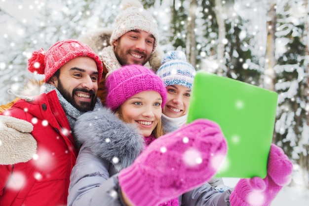 tecnología, temporada, amistad y concepto de la gente - grupo de hombres y mujeres sonrientes que toman selfie tablet pc en el bosque de invierno