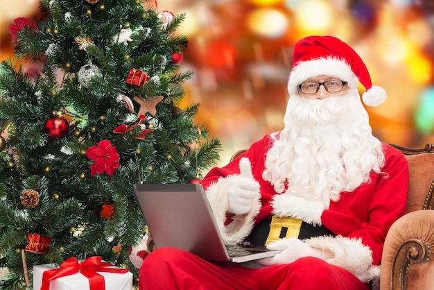 tecnología, vacaciones y concepto de personas - hombre disfrazado de santa claus con computadora portátil, regalos y árbol de navidad sentado en un sillón sobre fondo de luces rojas