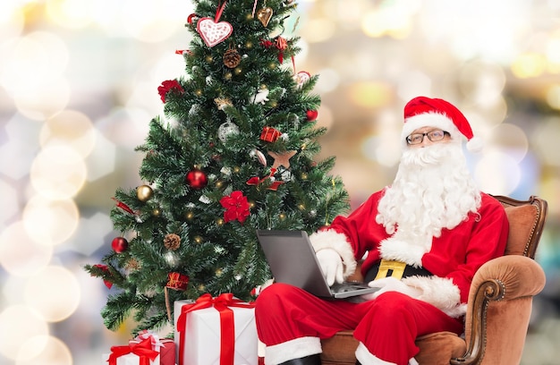 tecnología, vacaciones y concepto de personas - hombre disfrazado de santa claus con computadora portátil, regalos y árbol de navidad sentado en un sillón sobre fondo de luces