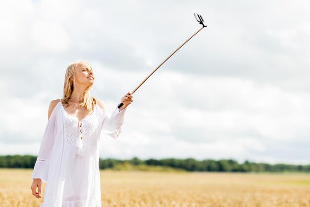 tecnología, vacaciones de verano, vacaciones y concepto de personas - mujer joven sonriente con vestido blanco tomando fotos con un palo selfie de smartphone en el campo de cereales