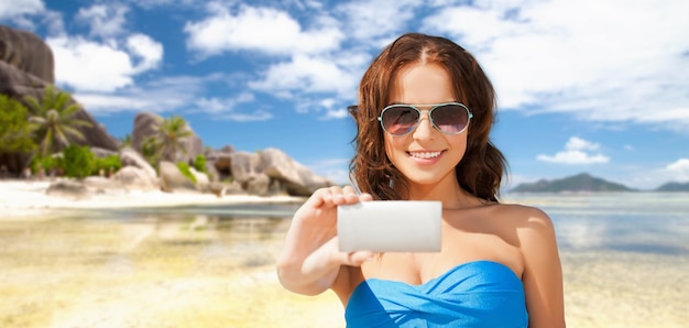 tecnología, vacaciones de verano, viajes y concepto de personas - mujer joven feliz en traje de baño bikini y gafas de sol tomándose selfie con smatphone sobre fondo de playa y palmeras