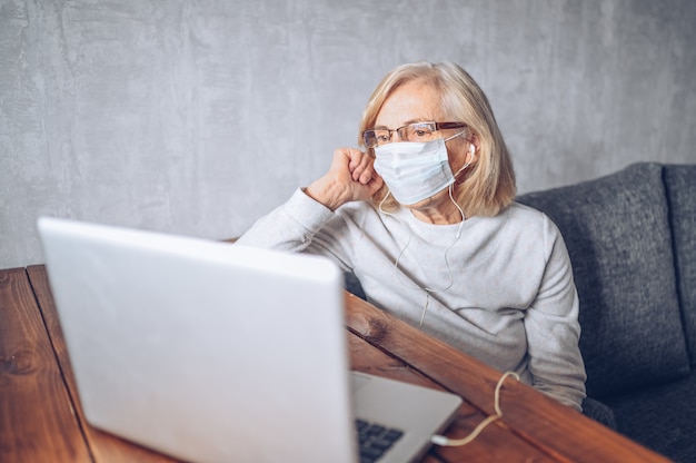 Tecnología, vejez y concepto de la gente - mujer mayor mayor triste sola en la máscara médica de la cara que trabaja y que hace una videollamada con la computadora portátil en casa durante la pandemia del coronavirus COVID19. Quedarse en casa