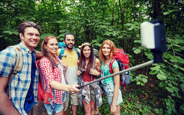 tecnologia, viagens, turismo, caminhada e conceito de pessoas - grupo de amigos sorridentes andando com mochilas tirando foto por smartphone na vara de selfie na floresta