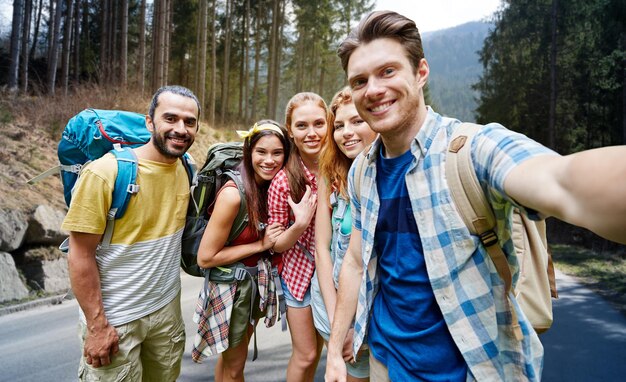 tecnologia, viagens, turismo, caminhada e conceito de pessoas - grupo de amigos sorridentes andando com mochilas tomando selfie por smartphone ou câmera sobre floresta e fundo de estrada