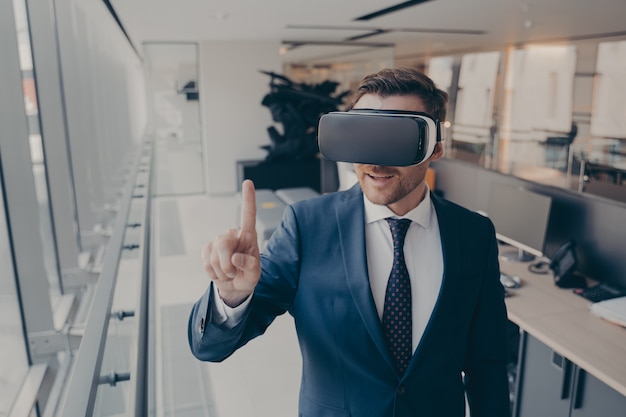 Tecnologias do futuro. Jovem empresário em um terno elegante usando fone de ouvido VR, tocando, explorando a realidade virtual no trabalho, tentando tocar em algo com o dedo indicador à sua frente, parado no escritório