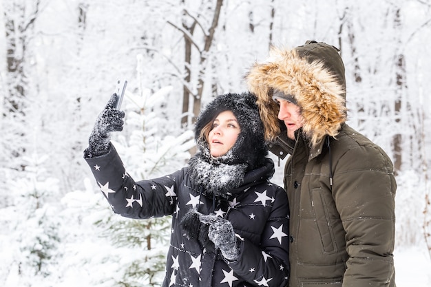 Tecnologias e conceito de relacionamento - casal feliz e sorridente tomando uma selfie em uma floresta de inverno do lado de fora.