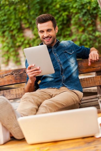 Las tecnologías facilitan la vida. Joven alegre sosteniendo tableta digital y sonriendo mientras está sentado en el banco de madera al aire libre