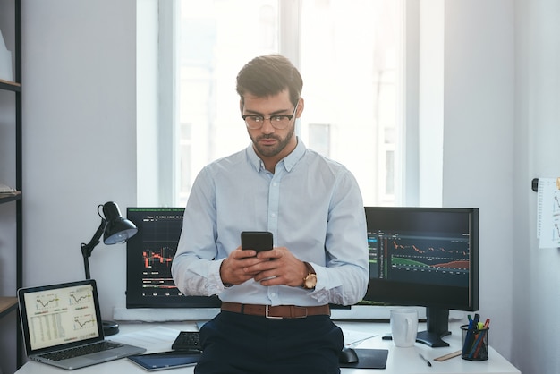 Las tecnologías modernas exitoso joven comerciante en ropa formal y anteojos con smartphone mientras