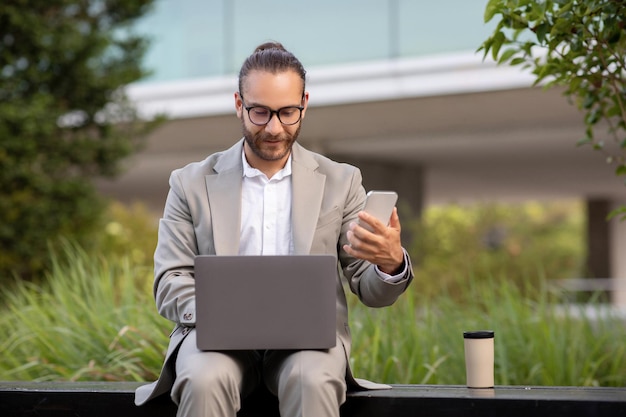 Tecnologías modernas para los negocios Joven empresario con portátil y teléfono inteligente sentado al aire libre