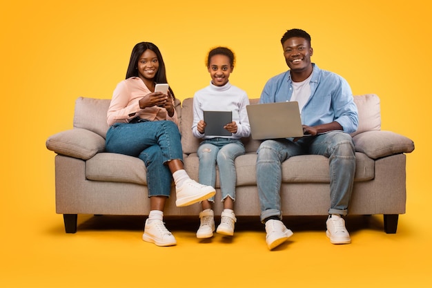 Tecnologias para a família Feliz mãe negra pai e sua filha sentada com diferentes gadgets fundo amarelo