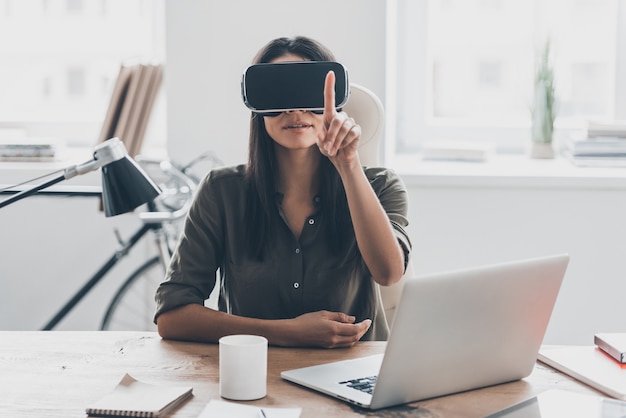 Tecnologías de realidad virtual. Mujer joven confiada en casco de realidad virtual apuntando en el aire mientras está sentada en su lugar de trabajo en la oficina