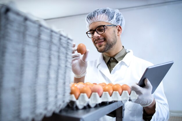 Tecnólogo de fábrica de alimentos con redecilla de bata blanca y guantes higiénicos que controlan la producción de huevos en la planta de procesamiento de alimentos en una tableta.