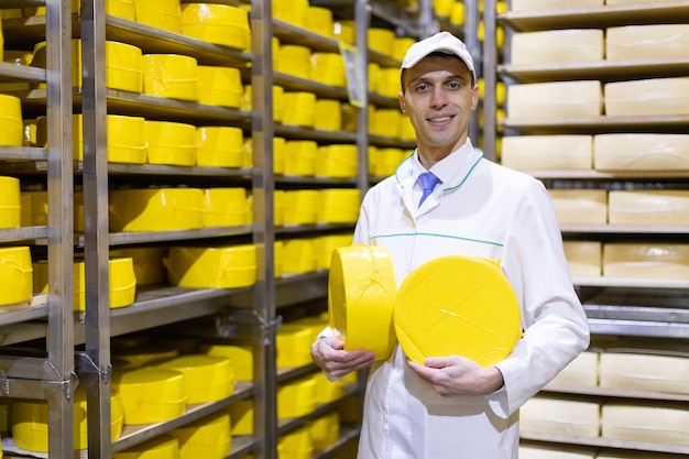 El tecnólogo de túnica blanca con una cabeza de queso amarillo en las manos está en la tienda para la producción de mantequilla y queso El proceso de producción en la planta de productos lácteos Bastidores con queso