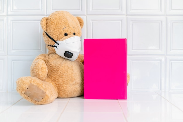 Teddybär mit Gesichtsmaske und rosa Notizbuch auf weißem Hintergrund.