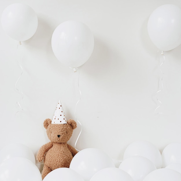 Teddybär mit Partyhut sitzt neben Luftballons vor weißem Hintergrund