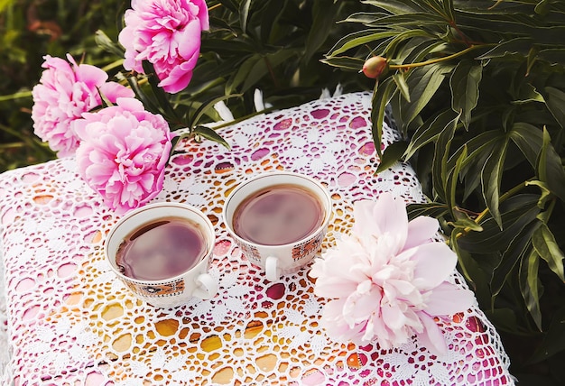 Tee im Landhausstil im Sommergarten Zwei Tassen schwarzer Tee auf handgefertigter gehäkelter Vintage-Lacy-Tischdecke und blühende Pfingstrosenblumen im Sonnenlicht