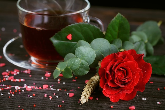 Tee in einer transparenten Tasse, Bonbons und eine rote Rose im Dunkeln