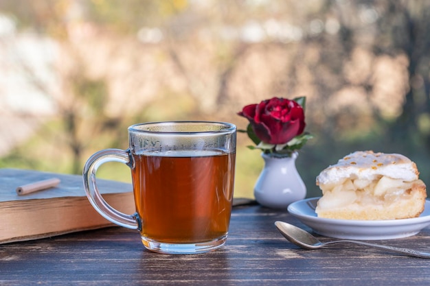 Teeglas mit süßem Kuchen und Buch auf Holztisch am Morgen im Freien aus nächster Nähe