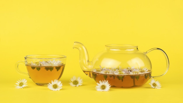 Teekanne und Glastasse mit Tee und Blumen auf gelbem Grund.