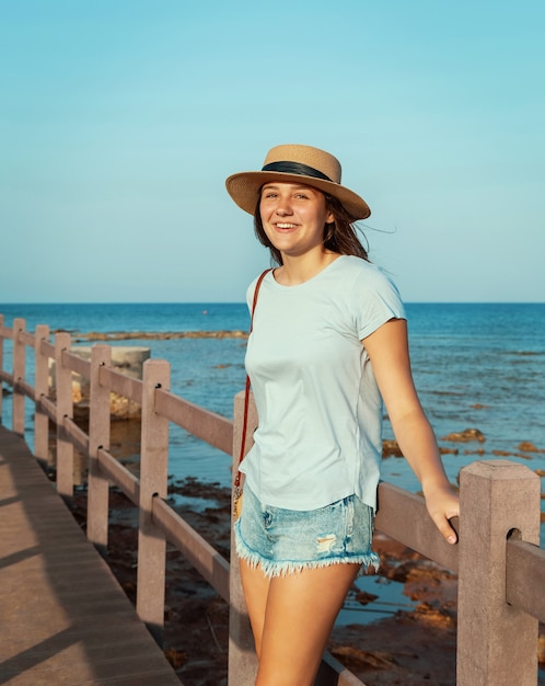 Teen Girl steht auf Holzsteg am Meer bei Sonnenuntergang, trägt hellblaues T-Shirt, Strohhut und Geldbörse. Sommerreisekonzept