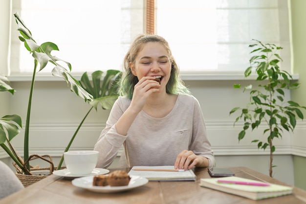 Teen Mädchen, das Cupcakes isst, Tee trinkt, zu Hause am Tisch sitzt