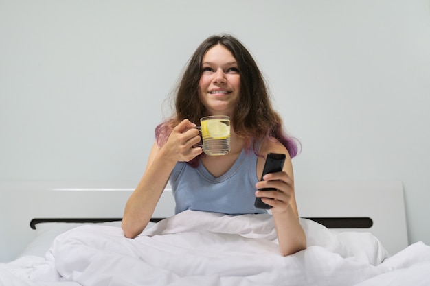 Teen Mädchen sitzt im Bett mit Glas Wasser mit Zitrone hält TV-Fernbedienung in der Hand