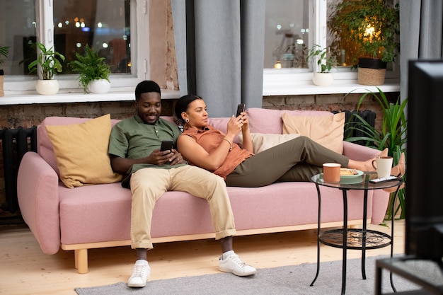 Teenager afrikanischer Abstammung und seine gemischtrassige Freundin mit Smartphones, die sich auf der Couch entspannen und in sozialen Netzwerken kommunizieren