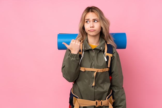 Teenager-Bergsteigermädchen mit einem großen Rucksack an der rosa Wand unglücklich und zur Seite zeigend