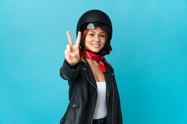 Teenager-Biker-Mädchen lokalisiert auf blauer Wand lächelnd und Siegeszeichen zeigend