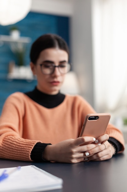 Teenager, der am Schreibtisch im Wohnzimmer sitzt, während er Universitäts-E-Mails auf einem modernen Smartphone überprüft. Schüler, die während der Coronavirus-Quarantäne Lifestyle-Informationen im Internet durchsuchen