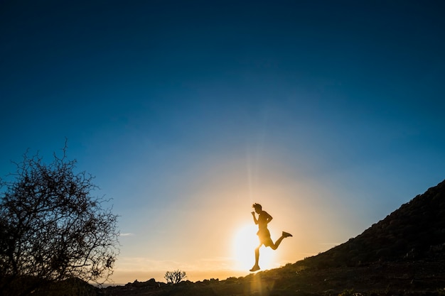 Teenager, der bei Sonnenuntergang in den Bergen läuft - Läuferleben und sportliches Konzept - Outdoor-Lifestyle - Baum und blauer Hintergrund