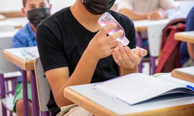 Teenager in der Schule, der seine Hände sterilisiert, während er eine Gesichtsmaske trägt, um eine Infektion zu verhindern