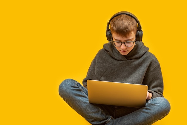 Teenager in Jeans, Kapuzenpullover, Brille und Kopfhörer sitzt mit gekreuzten Beinen und hält einen Laptop. Online-Lernen, Programmieren für Kinder, Kopierraum