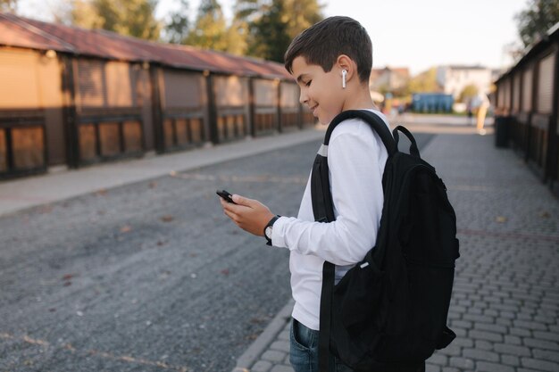 Teenager in weißem Sweatshirt und mit schwarzem Backpuck verwenden Telefon und kabellose Kopfhörer und drucken Nachricht im sozialen Netzwerk