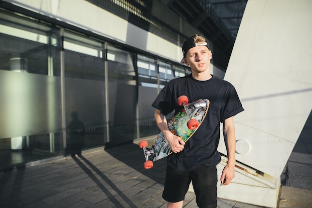 Teenager ist ein Skater in einer Mütze und mit einem Brett in den Händen, das im Hintergrund eines modernen Gebäudes spaziert.