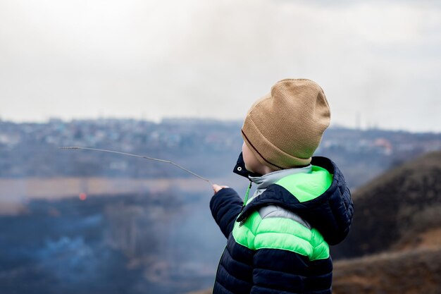 Teenager-Junge mit beigem Hut und Jacke auf dem Hintergrund von verbranntem Schilf, Bäumen, rauchiger Atmosphäre