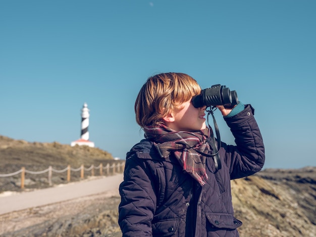 Teenager-Junge trägt warme Jacke und Wollschal und blickt an einem sonnigen Tag auf ein Fernglas, das am felsigen Ufer des Meeres sitzt