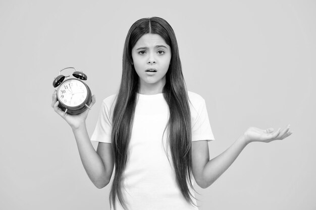 Teenager-Kind hält die Uhr auf gelbem Studio-Hintergrund isoliert Teenager-Kind mit Wecker zeigt die Zeit Wütendes Gesicht verärgert Emotionen von Teenager-Mädchen